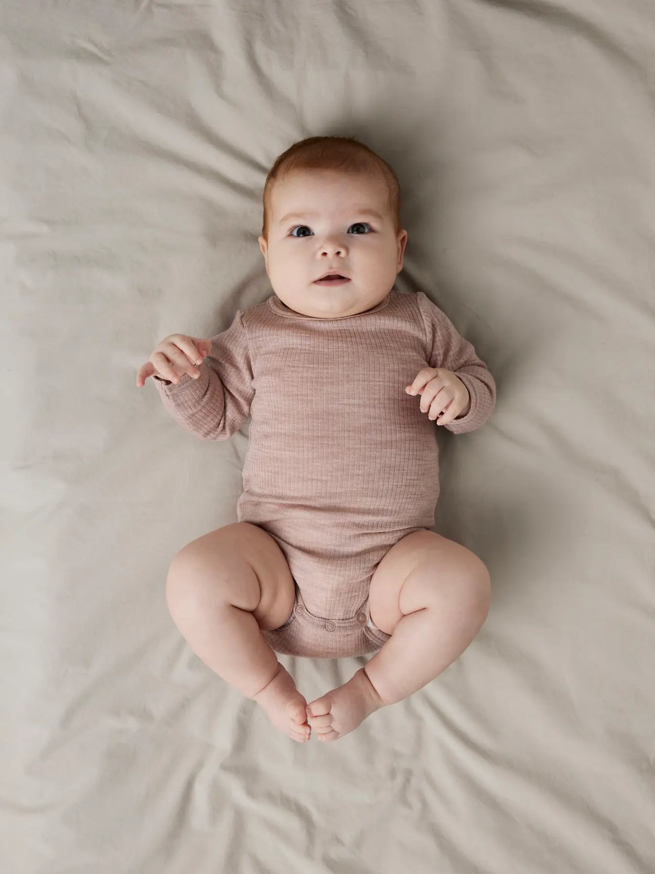 Baby trägt altrosafarbenen Langarmbody aus Merinowolle auf hellem Untergrund.