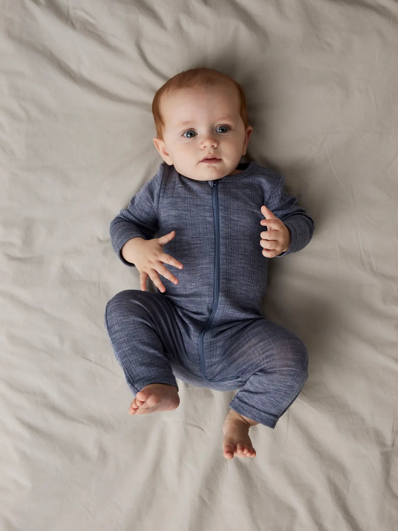 Ein Baby liegt in einem blauen Merino-Schlafanzug von Name It auf dem Bett.