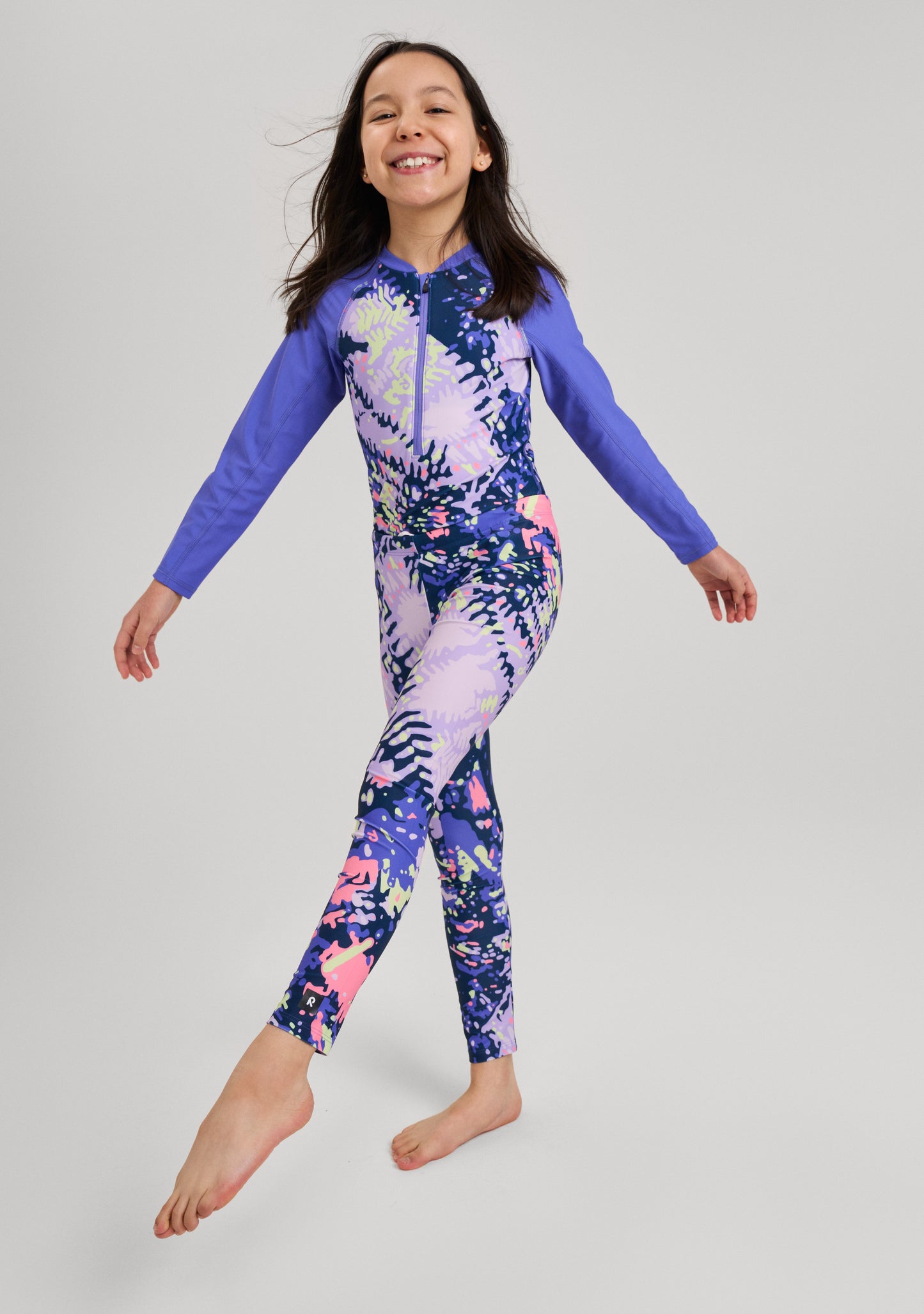 Mädchen trägt den lila Reima Badeanzug Aallot mit floralem Muster und langen Ärmeln; die gezeigte Leggings gehört nicht zum Lieferumfang