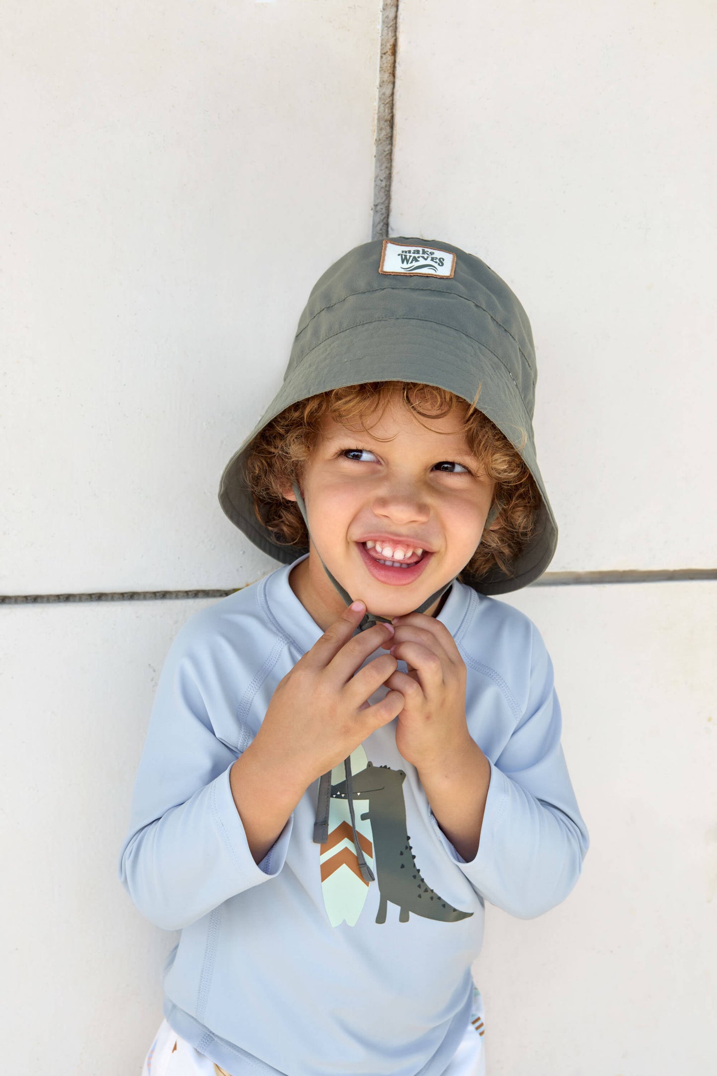 Kind mit LÄSSIG UV-Shirt und Sonnenhut steht an Wand – sicher & stilvoll mit Bademode von Bärechind in Winterthur, Zürich, Schweiz.