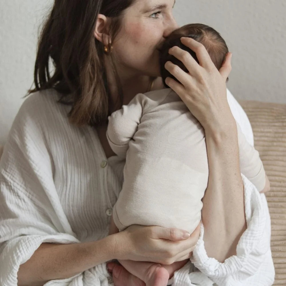 Baby im beigen Emma & Noah Wickelbody aus Merinowolle auf dem Arm seiner Mutter