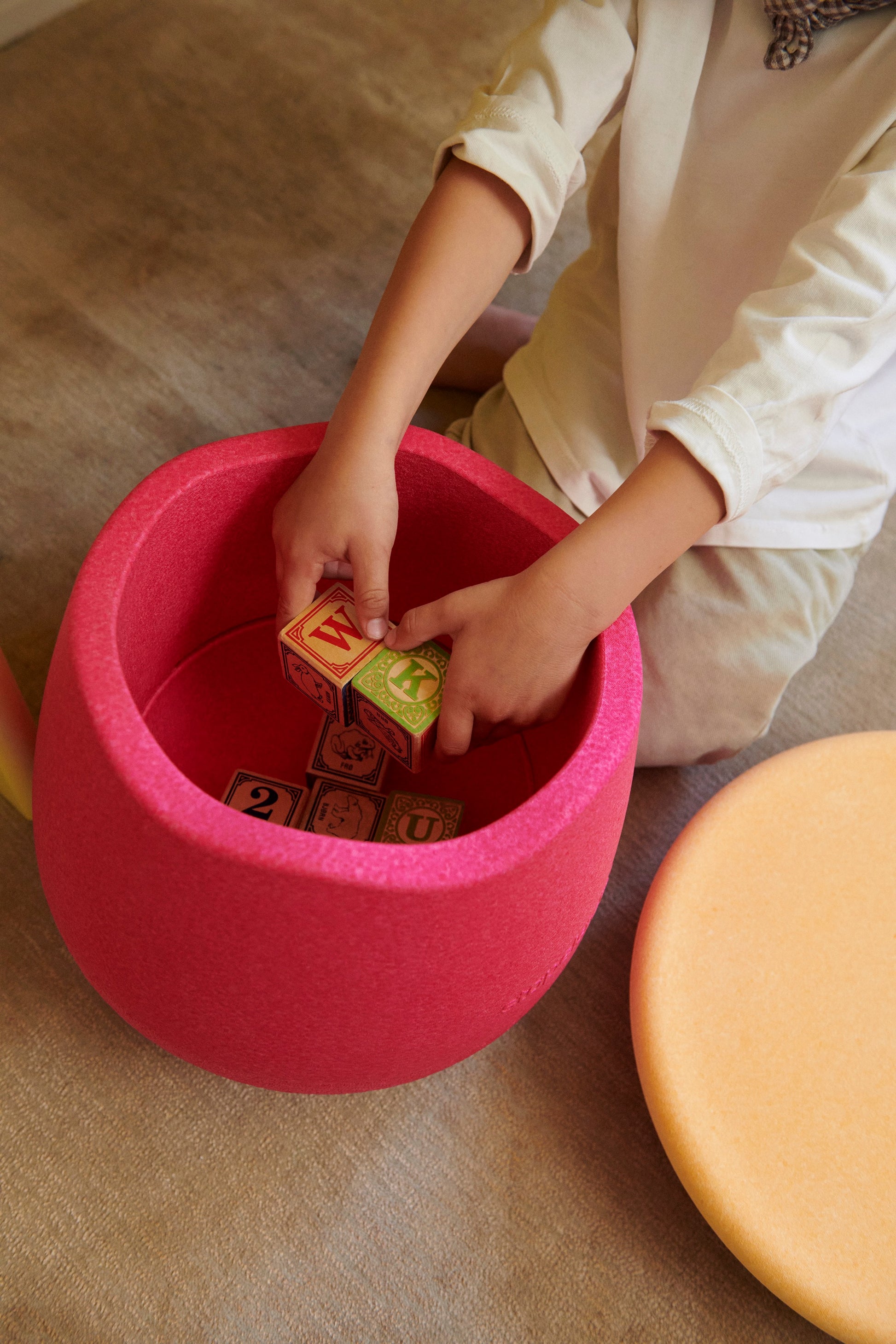 Kind legt Spielzeug in einen pinken Schaumstoff-Sitzhocker mit Stauraum.