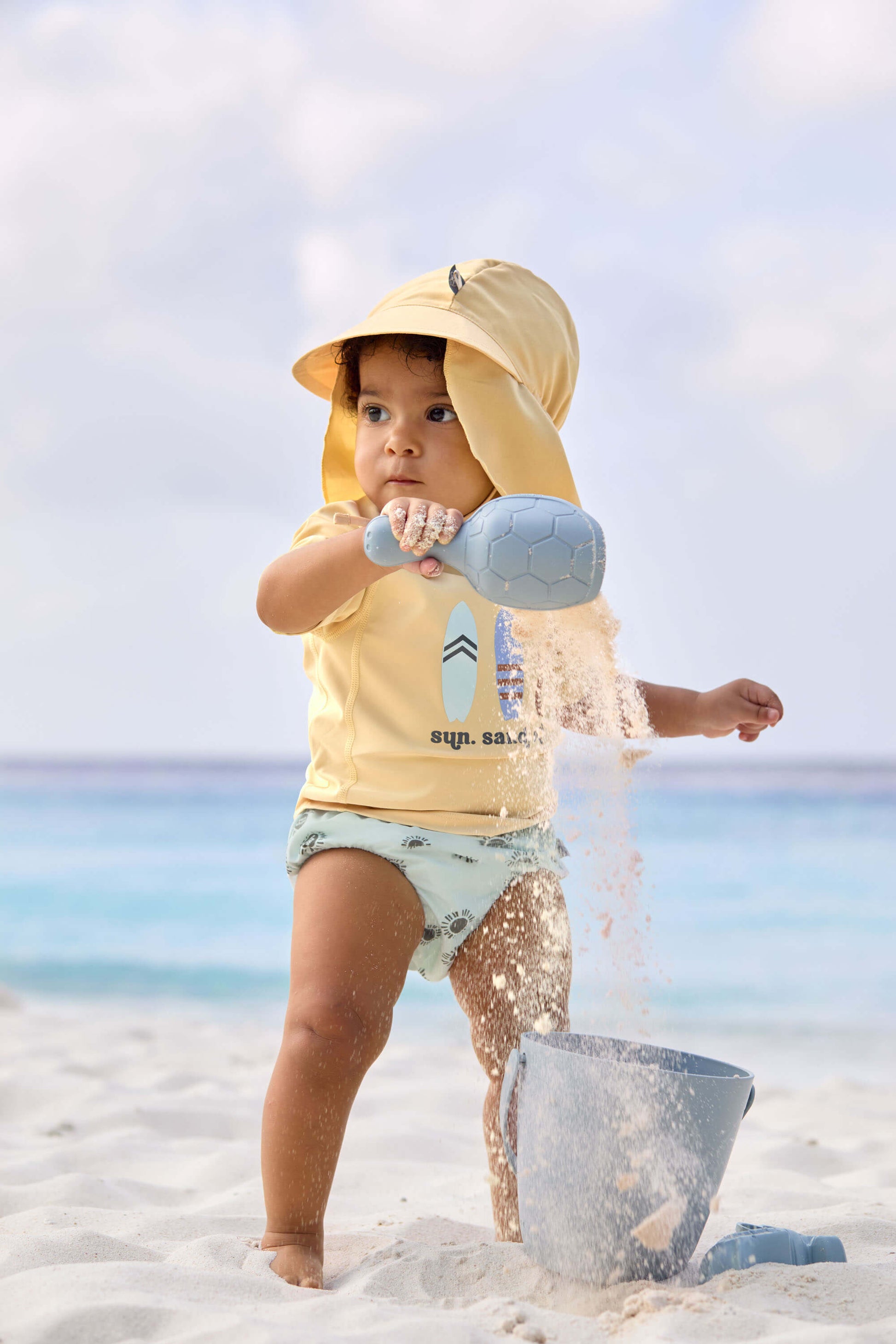 Sonnenhut von Lässig für Babys in Gelb – mit UV-Schutz 80 & langem Nackenschutz, Angezogen an Strand.