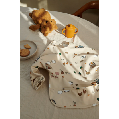 Kind trägt das Liewood Merle Langarm-Lätzchen mit Bauernhof-Print beim Essen am Tisch