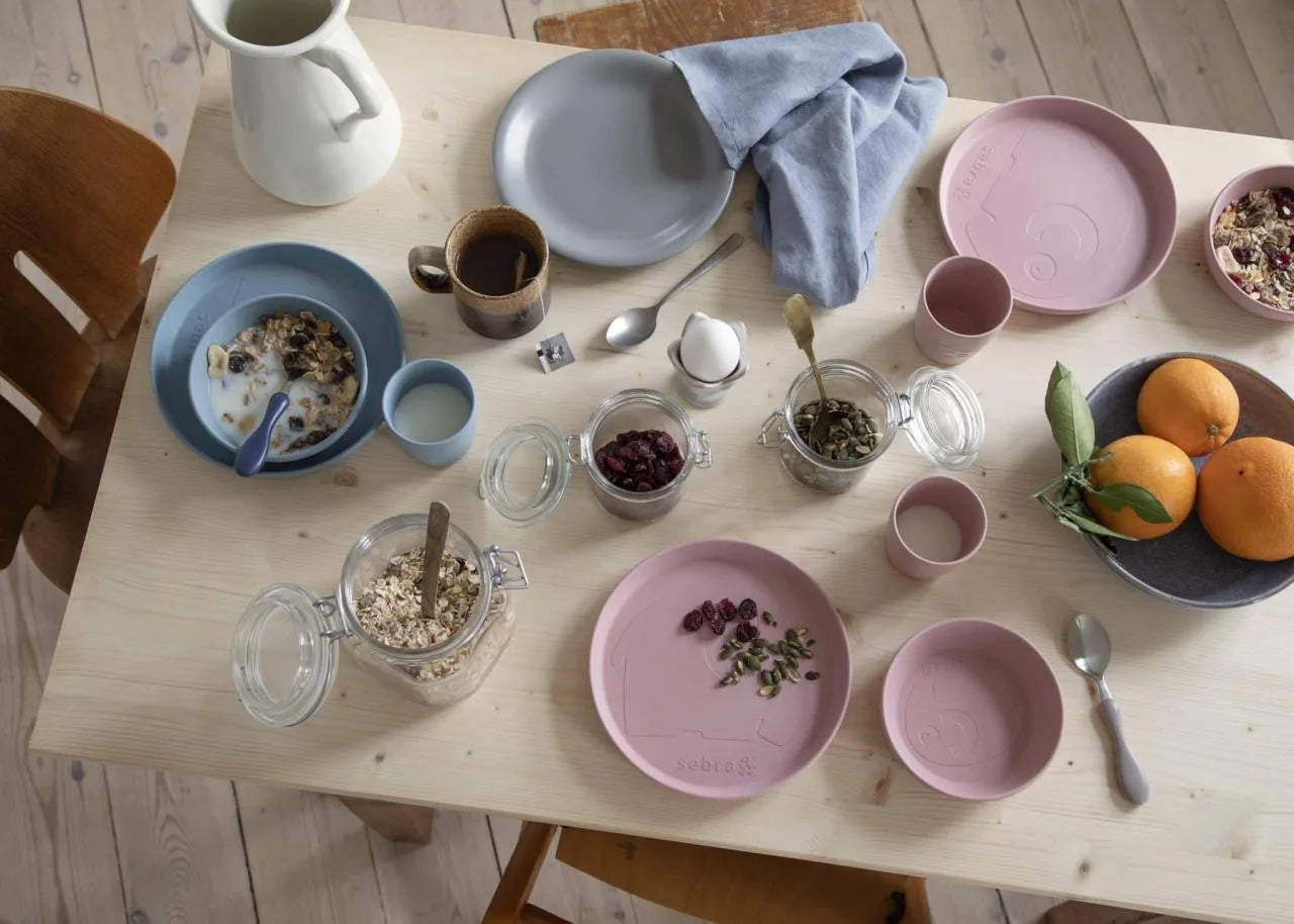 Rosa MUMS-Essset auf einem hell gedeckten Familientisch zwischen Obst, Beeren und Geschirr arrangiert.