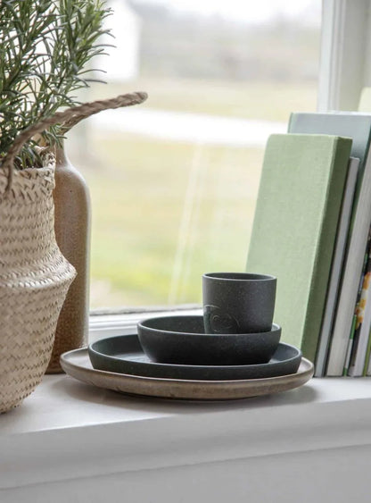 Dunkelgrünes MUMS-Geschirrset aus Teller, Schale und Becher, dekorativ auf einem Tisch am Fenster arrangiert.