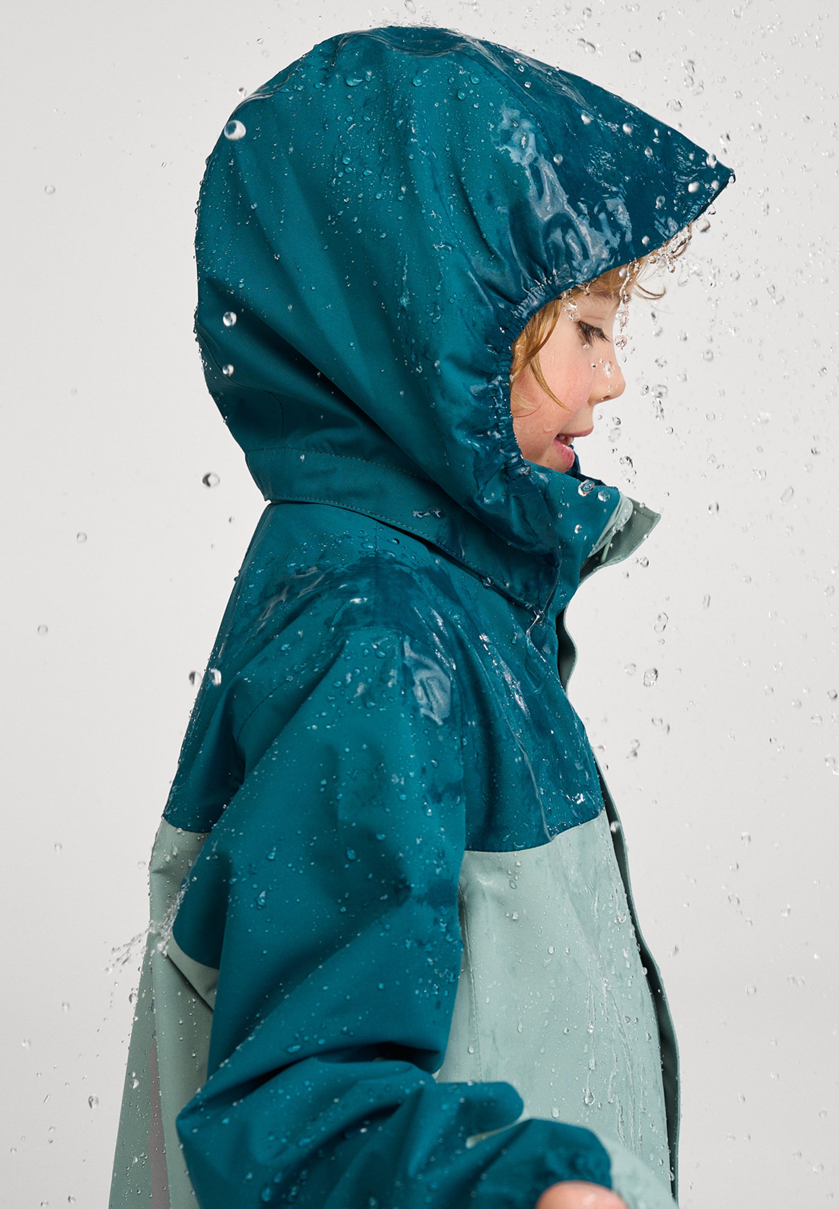 Kind seitlich trägt ReimaTec Regenjacke Salmi in Stone Green bei Regenwetter